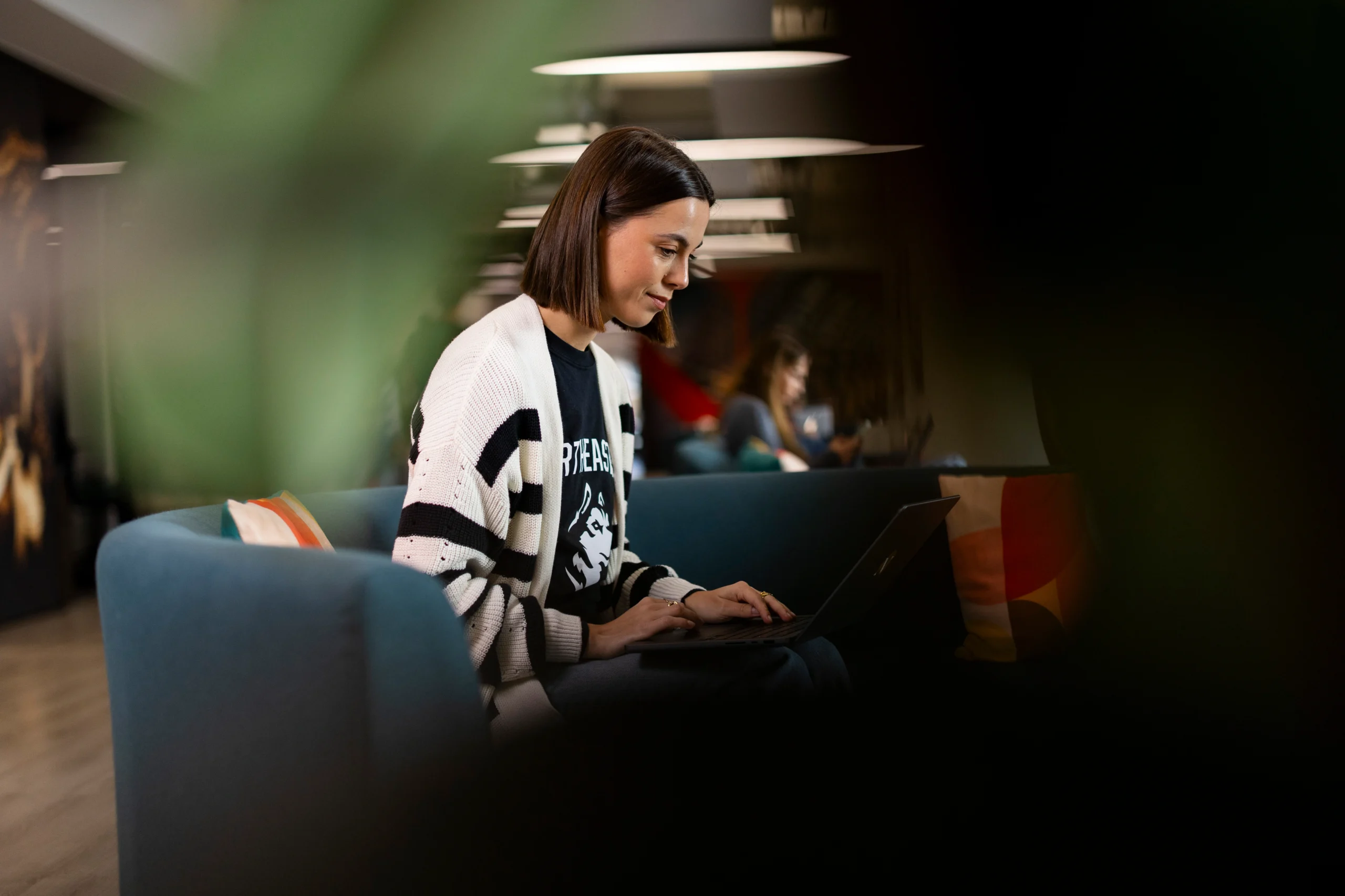 A woman with a bob haircut sits on a blue couch, typing on a laptop. She is wearing a black and white striped sweater. The background is softly blurred, with another person visible farther back.