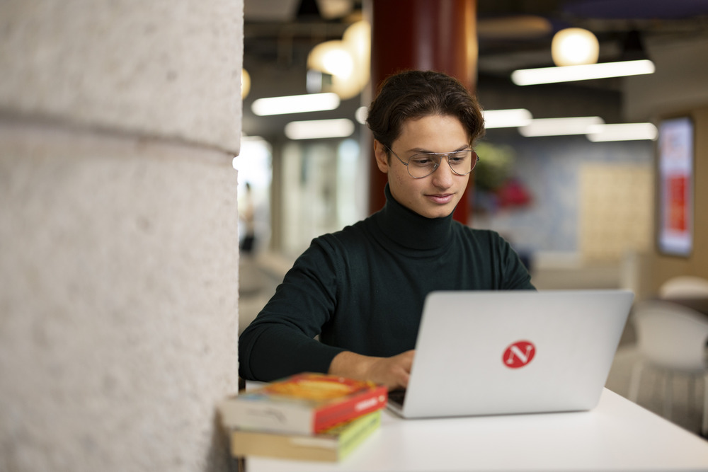 A person wearing glasses and a dark turtleneck works on a laptop with a red “N” sticker, sitting at a table with books in a modern, well-lit indoor space.