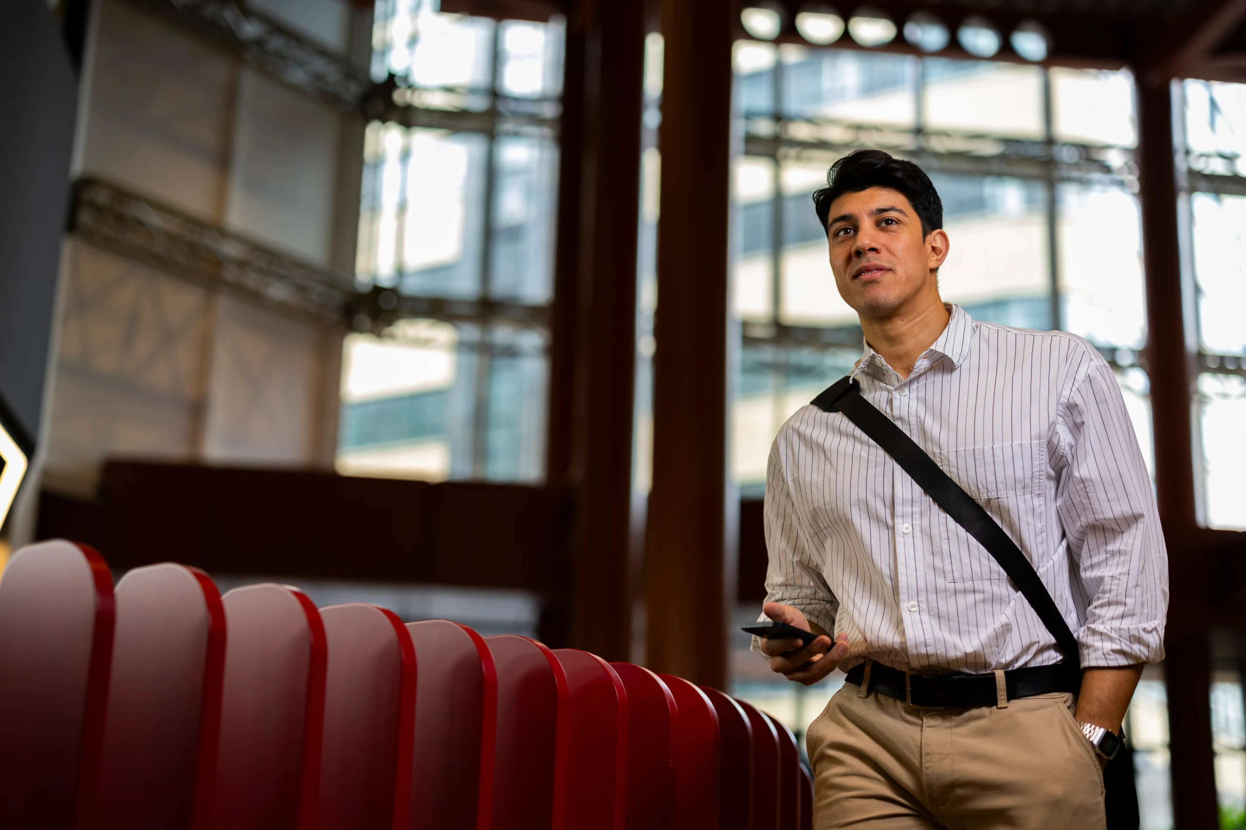 A man in a striped shirt stands indoors near a red barrier, holding a phone and wearing a shoulder bag, with large windows and modern architecture in the background.