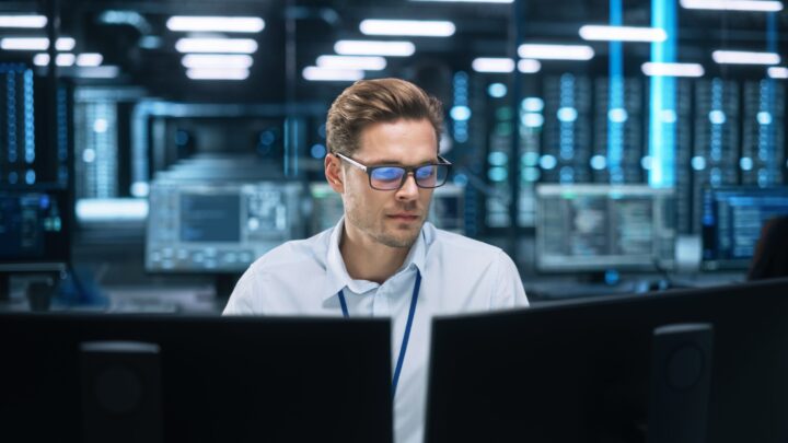 A man wearing glasses and a white shirt works at a desk with multiple monitors in a modern, high-tech office with servers and computer screens in the background.