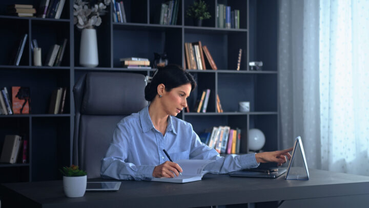 A woman in a blue shirt sits at a desk, writing in a notebook while using a tablet. She is in a modern office with a bookshelf and various items in the background.