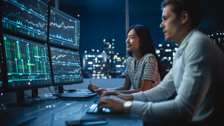 Two people sit at a desk in an office at night, looking at multiple monitors displaying graphs and financial data, with city lights visible through large windows in the background.