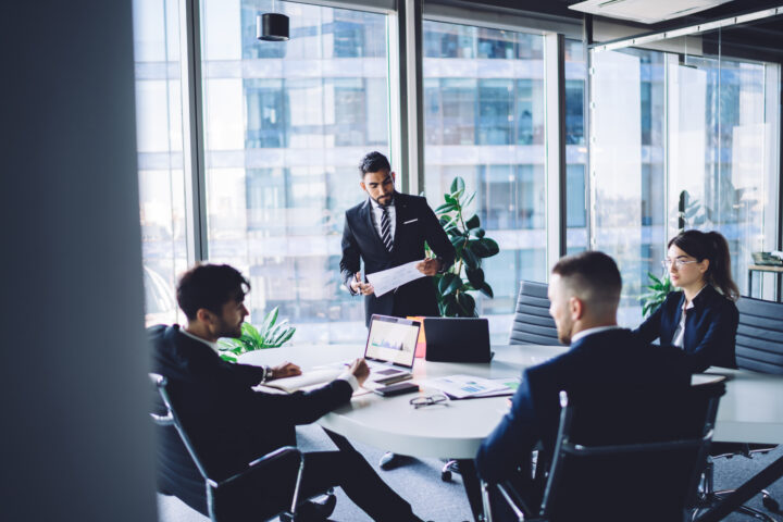 Four business professionals in formal attire are having a meeting in a modern glass-walled office. One person is standing and speaking while holding papers, and three others are seated at a round table with laptops and documents.