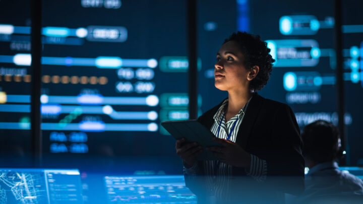 A woman holding a tablet stands in a modern control room, illuminated by blue screens displaying data and digital graphics, suggesting a high-tech or cybersecurity environment.