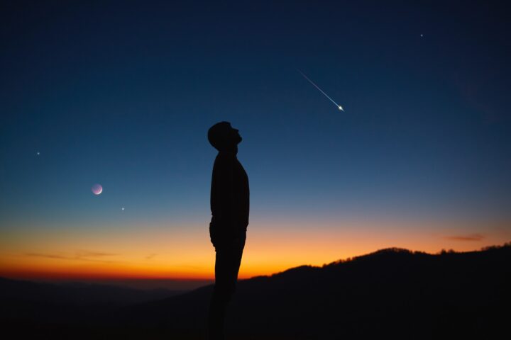 A silhouette of a person looking up at the night sky with a meteor streaking by, planets visible, and a colorful sunset behind distant hills.