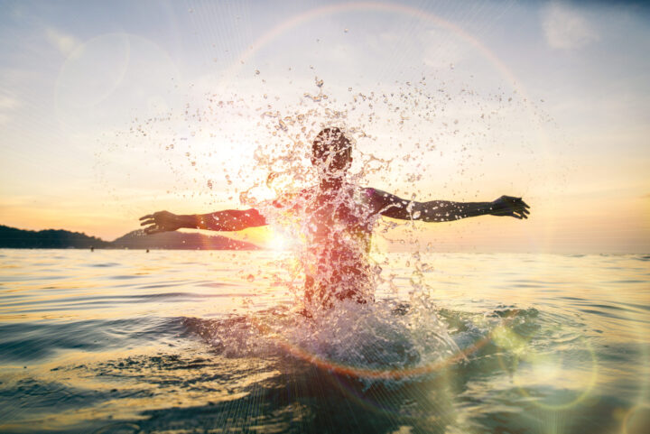A person stands in the ocean, arms outstretched, splashing water with sunlight shining behind them. The scene is bright and energetic, with sun rays and lens flares enhancing the sunset atmosphere.