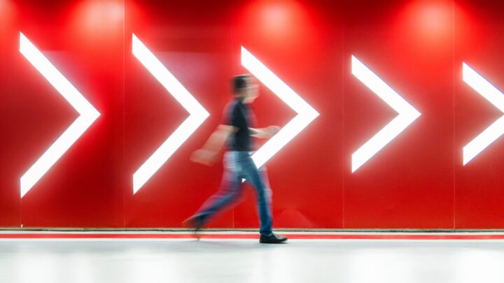 A blurred person walks past a bright red wall decorated with large white arrow symbols pointing to the right, passing signage for an Apprenticeship funding event.