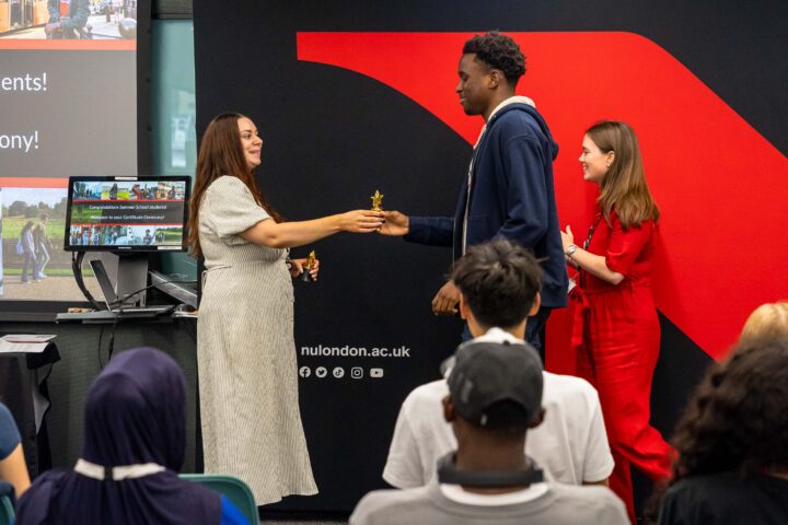 A woman hands a small trophy to a smiling young man on stage, while another woman in red stands beside them. Several people sit and watch, and a large red and black backdrop is behind them.