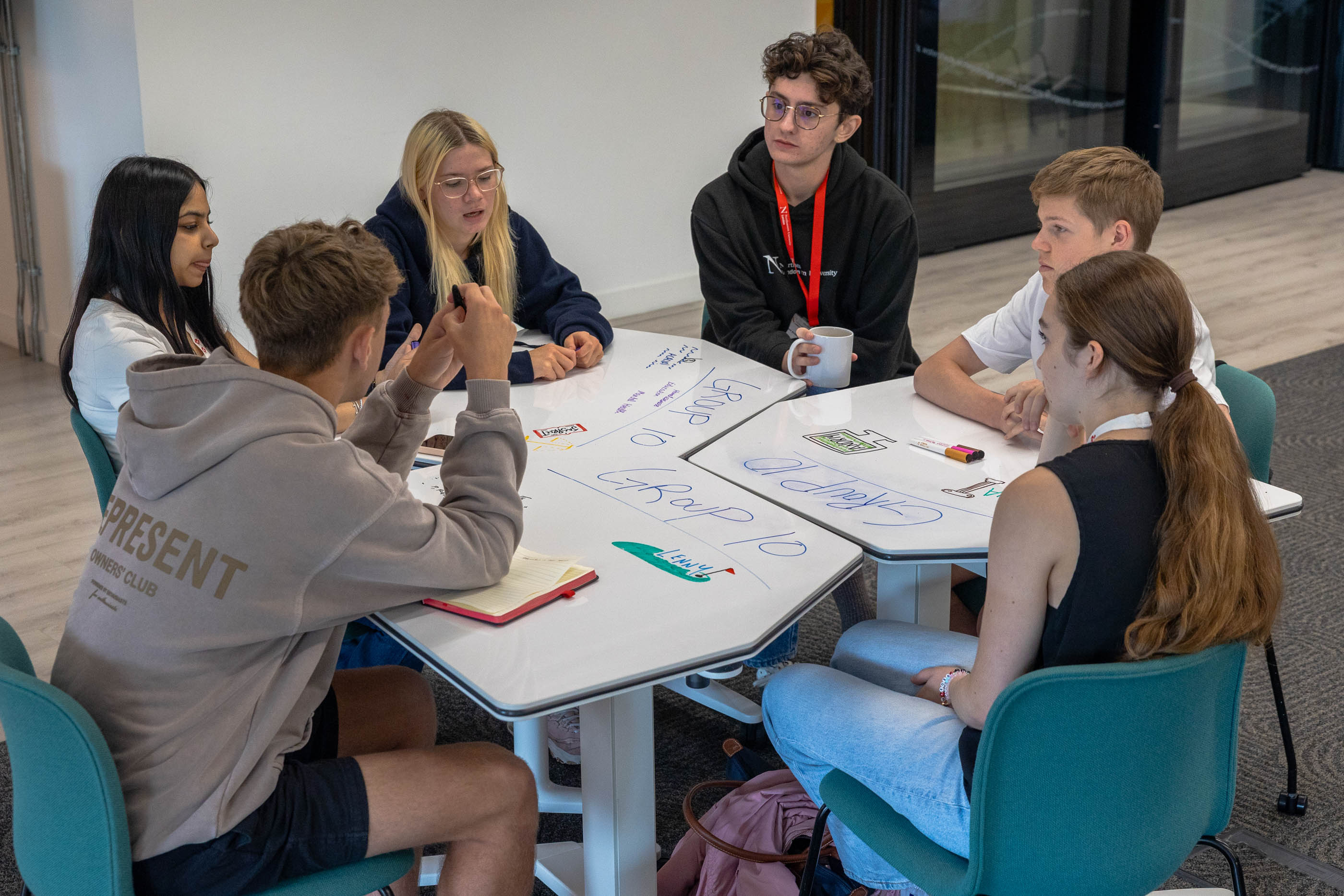 Six young adults sit around a table covered with notes and markers, engaged in discussion. Some are writing or holding papers, and one has a coffee mug. They appear to be collaborating in a modern workspace.