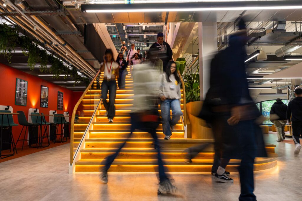 People walk up and down a wide, yellow-lit staircase in a modern office space with red walls, bar seating, and exposed ceilings. The movement creates blurred figures, conveying a busy, dynamic atmosphere.