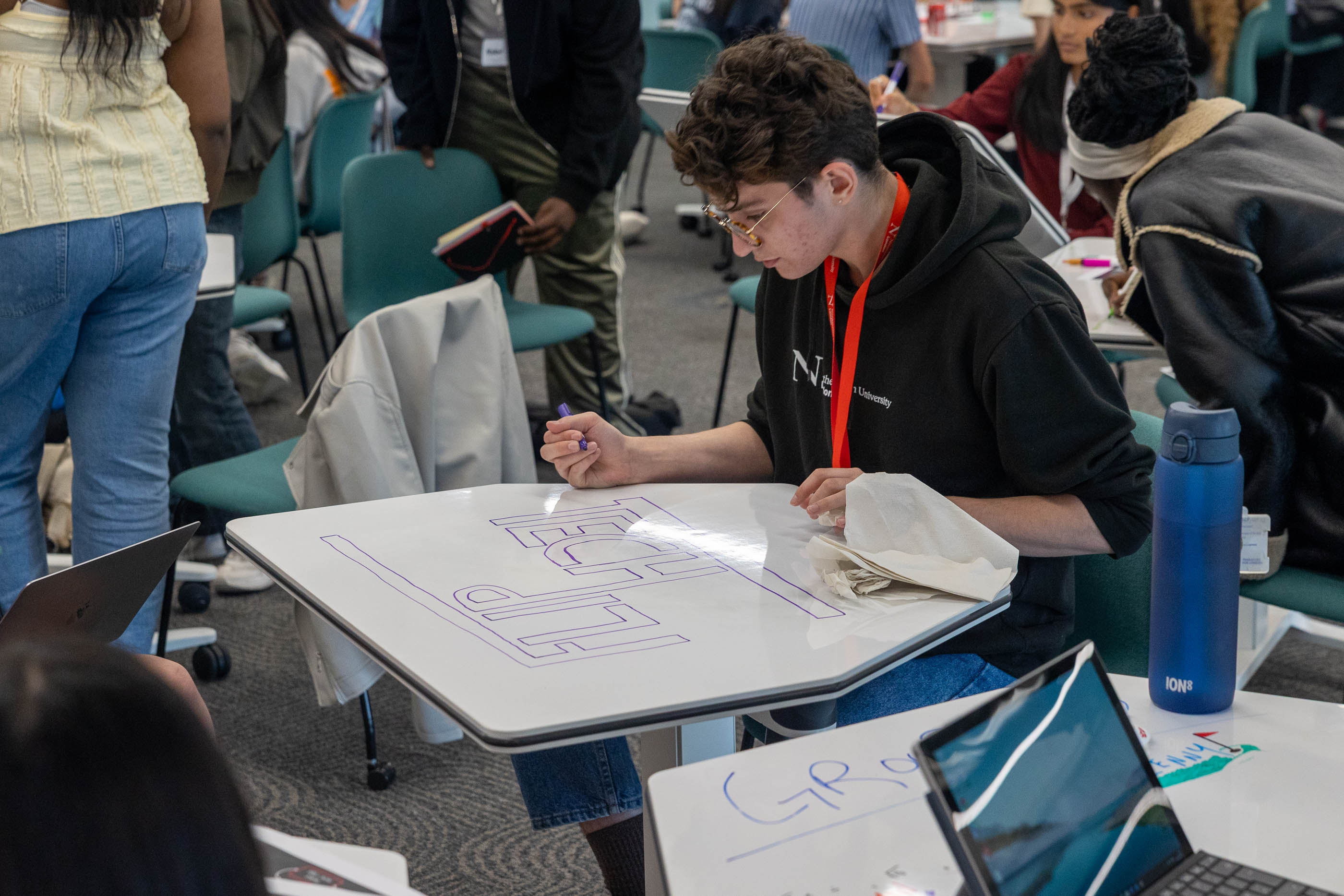 A person with short hair and glasses draws large block letters on a whiteboard table in a classroom, surrounded by other students and laptops.