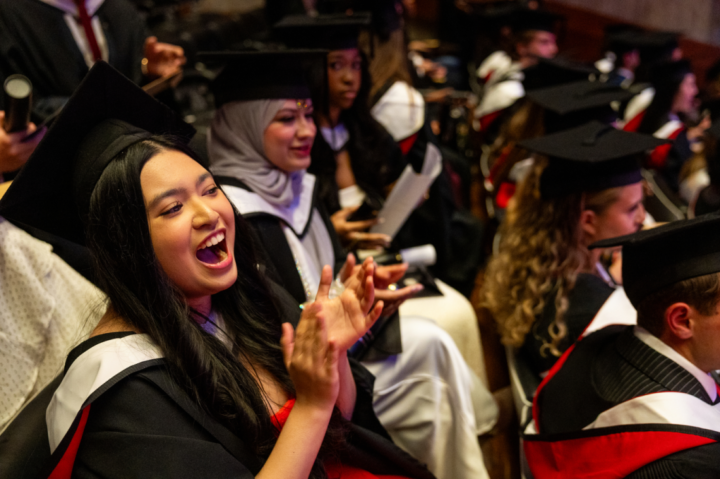 Graduates in caps and gowns sit in rows at a ceremony. A woman in the foreground is smiling and clapping enthusiastically, while others around her listen and hold their diplomas.
