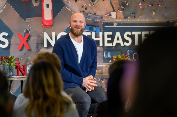 A man with a beard and blue sweater sits smiling onstage in front of an audience, with large red and white text and an aerial city street scene in the background.