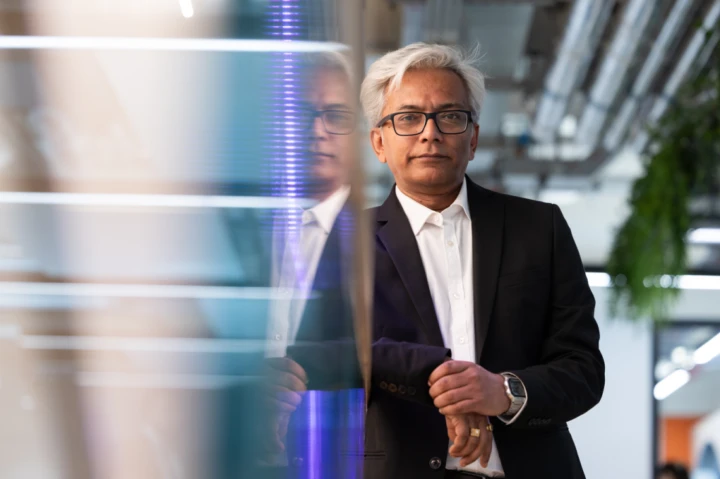 A man with gray hair and glasses, dressed in a black suit and white shirt, stands indoors with his reflection visible on a glass surface next to him. Industrial pipes and hanging plants are in the background.