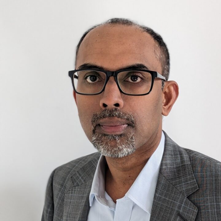 Bipin Rajendran, a man with short hair, a mustache, and a beard wearing glasses, a gray checked blazer, and a white shirt, stands against a plain white background.