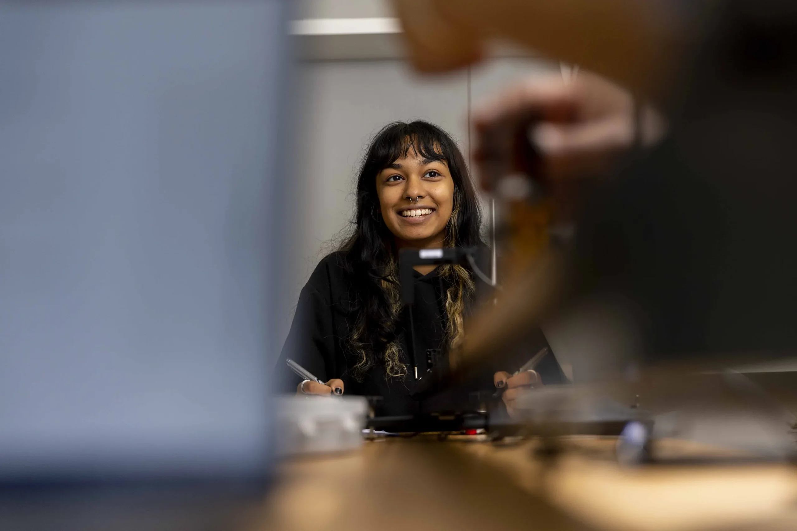A woman with long dark hair and bangs smiles while sitting at a desk, holding a pen—perhaps ready for her Business BSc studies. Her face is in focus, while objects and a person's hand in the foreground are blurred.