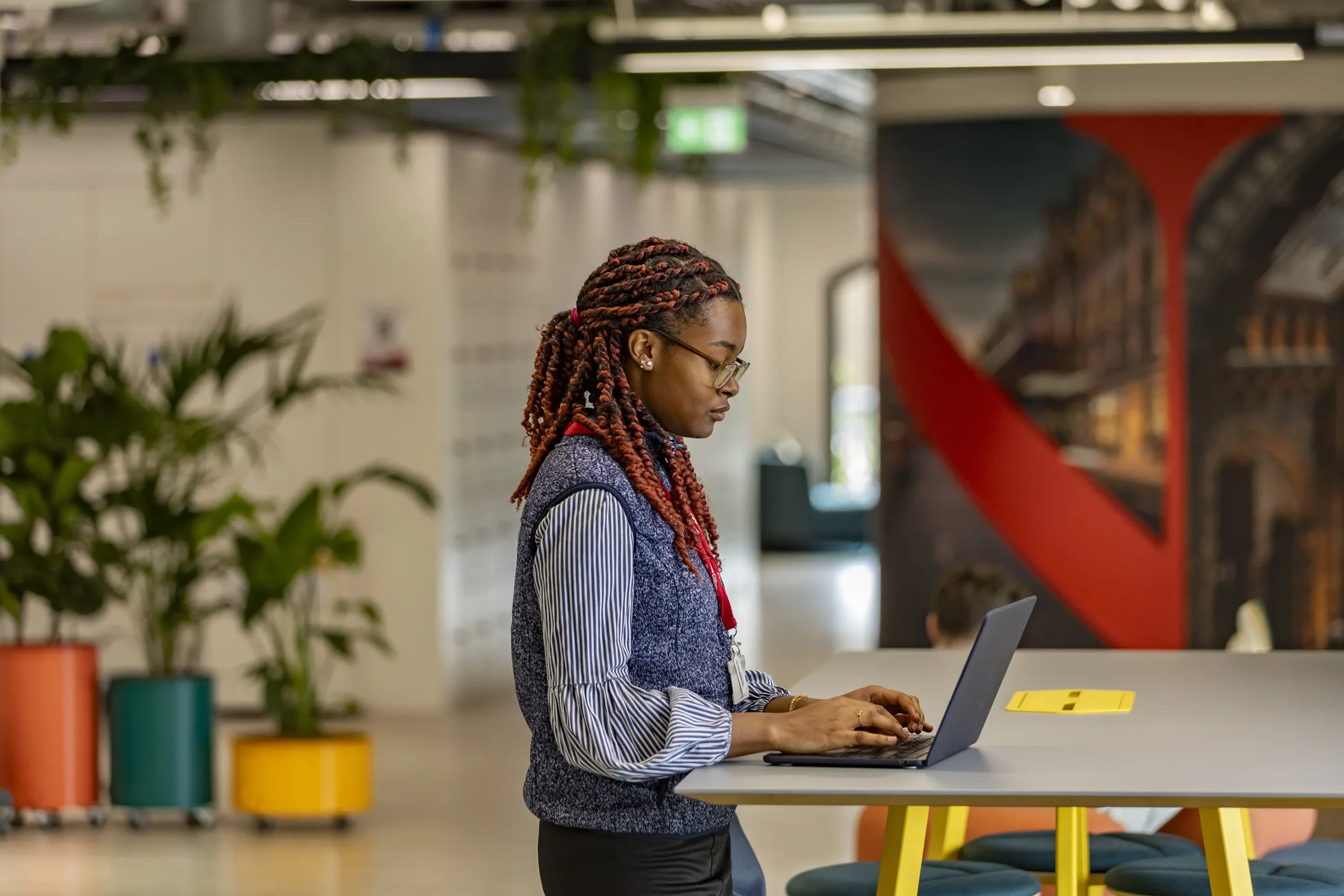 A woman with braided hair and glasses types on a laptop at a high table in a modern, colorful office space with plants and bright decor, working on her Business BSc studies.