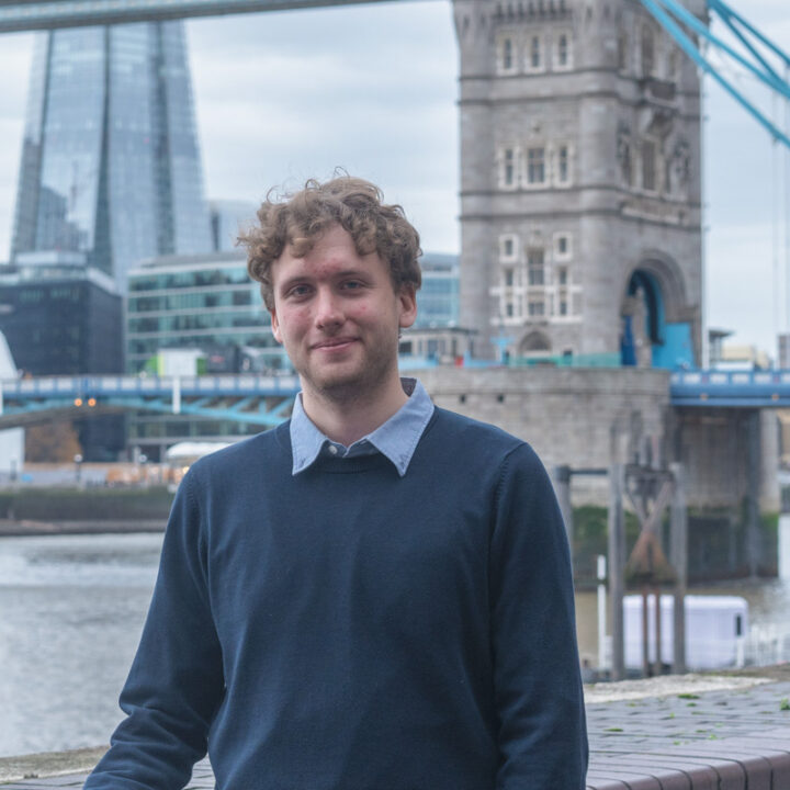 A man with curly hair and a navy sweater stands by the river with London's Tower Bridge and modern city buildings visible in the background.