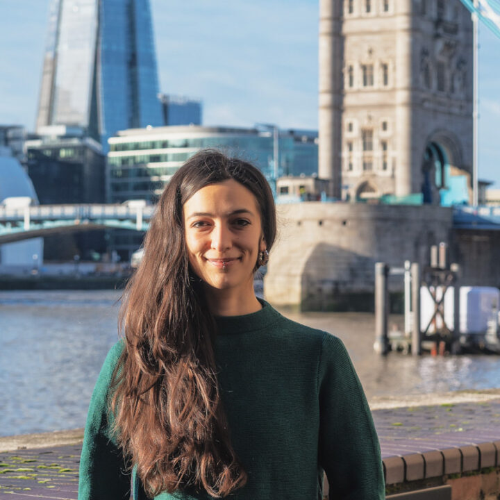 A woman with long dark hair, wearing a green sweater, stands smiling by a river with modern and historic buildings, including London’s Tower Bridge, in the background on a sunny day.
