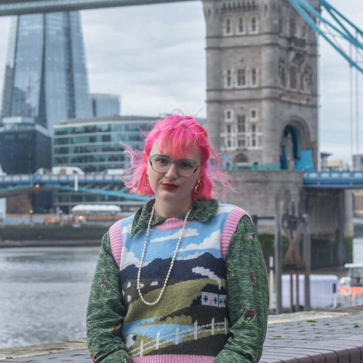 A person with bright pink hair, glasses, and red lipstick stands by the river in front of Tower Bridge in London, wearing a colorful sweater with a landscape design and a long pearl necklace.