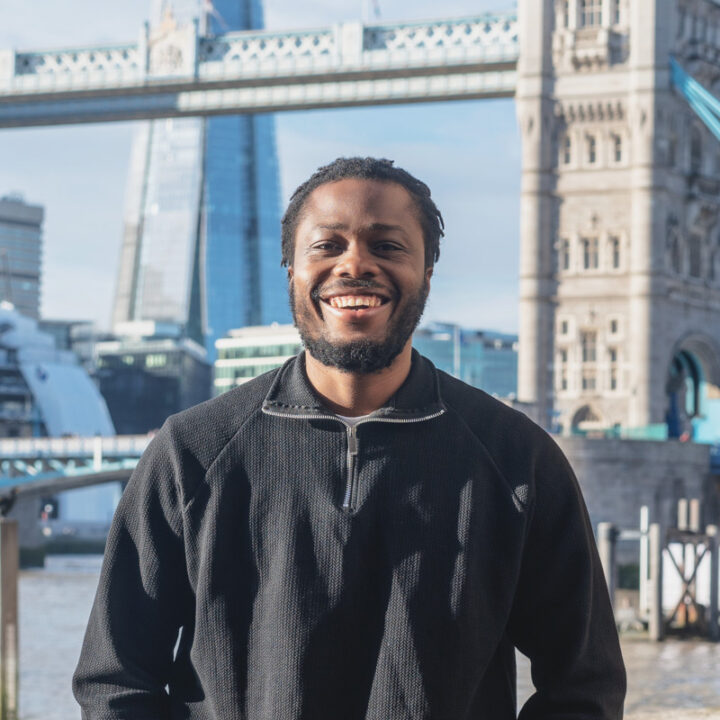 A smiling man with a beard and black sweater stands outdoors by the River Thames, with Tower Bridge and The Shard skyscraper visible in the background on a sunny day in London.