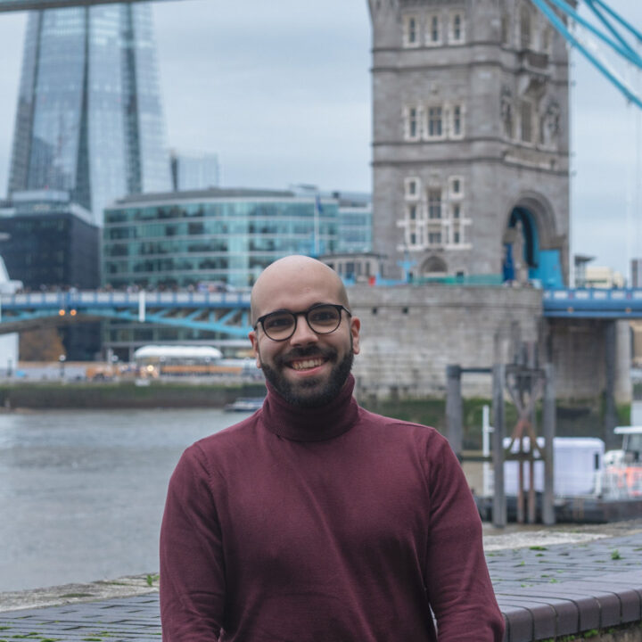 A smiling person wearing glasses and a maroon turtleneck stands by a riverside with London’s Tower Bridge and modern city buildings in the background.