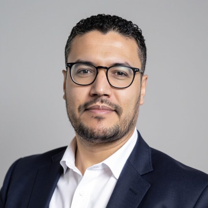 Houssem Sifaou, a man with short curly dark hair, a trimmed beard, and glasses, wearing a dark suit jacket and white shirt, poses against a plain light gray background.