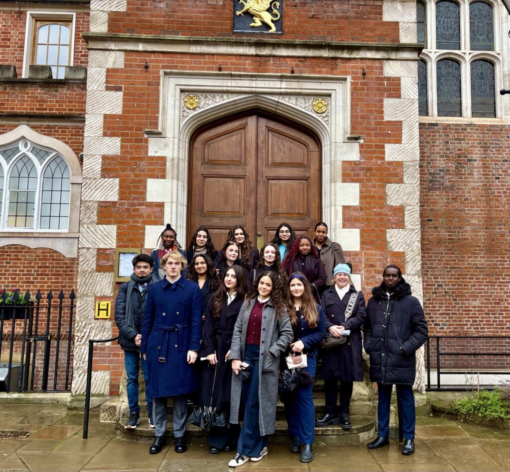 A group of 16 people, mostly young adults, stand together outside a historic brick building with large wooden doors and arched windows, posing for a group photo on a cloudy day.