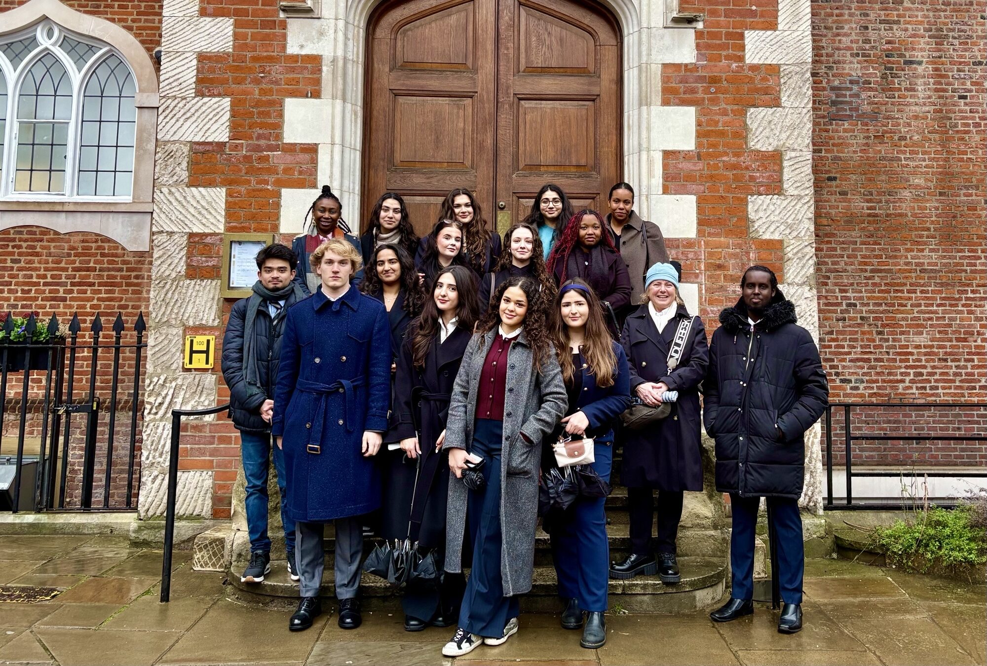 A group of 16 people, mostly young adults, stand together outside a historic brick building with large wooden doors and arched windows, posing for a group photo on a cloudy day.