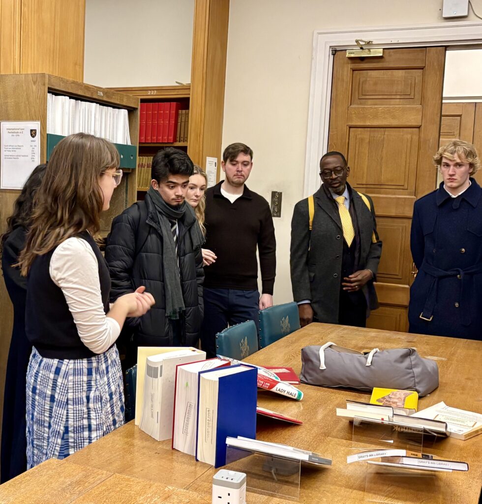 A group of six people stands around a table in a library or study room, listening to a woman speak. The table is covered with books, documents, and a gray backpack. Shelves and a wooden door are in the background.