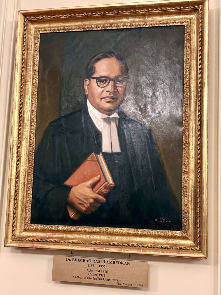 A framed portrait painting of Dr. Bhimrao Ramji Ambedkar in formal attire, holding a book. A plaque below identifies him as the author of the Indian Constitution and gives details about his life and achievements.