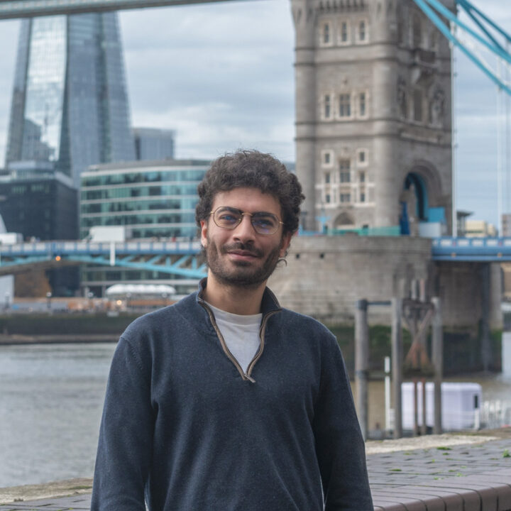 A man with curly hair and glasses stands near a river with the Tower Bridge and modern buildings in the background on a cloudy day.
