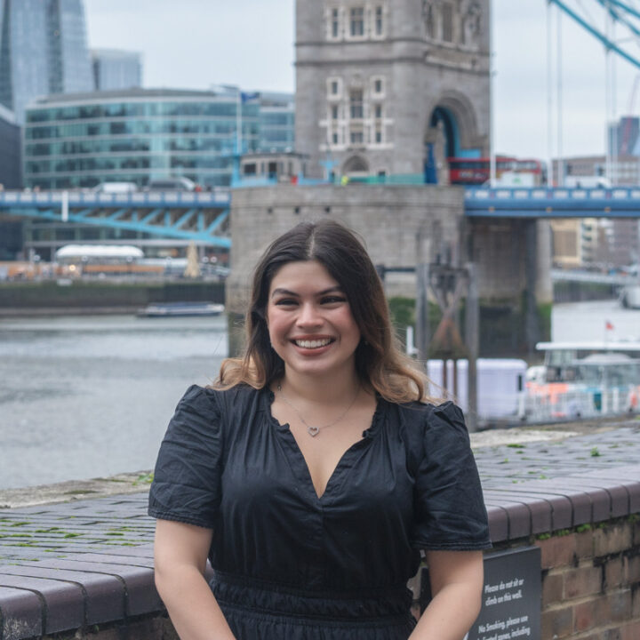 A young woman with long brown hair, wearing a black dress, smiles while standing in front of the Thames River and Tower Bridge in London on a cloudy day.