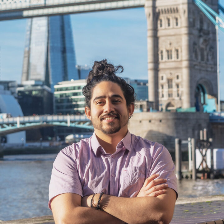 A man with long dark hair in a bun, wearing a light purple shirt, stands with arms crossed and smiles by the river. Tower Bridge and modern buildings are visible in the sunny background.