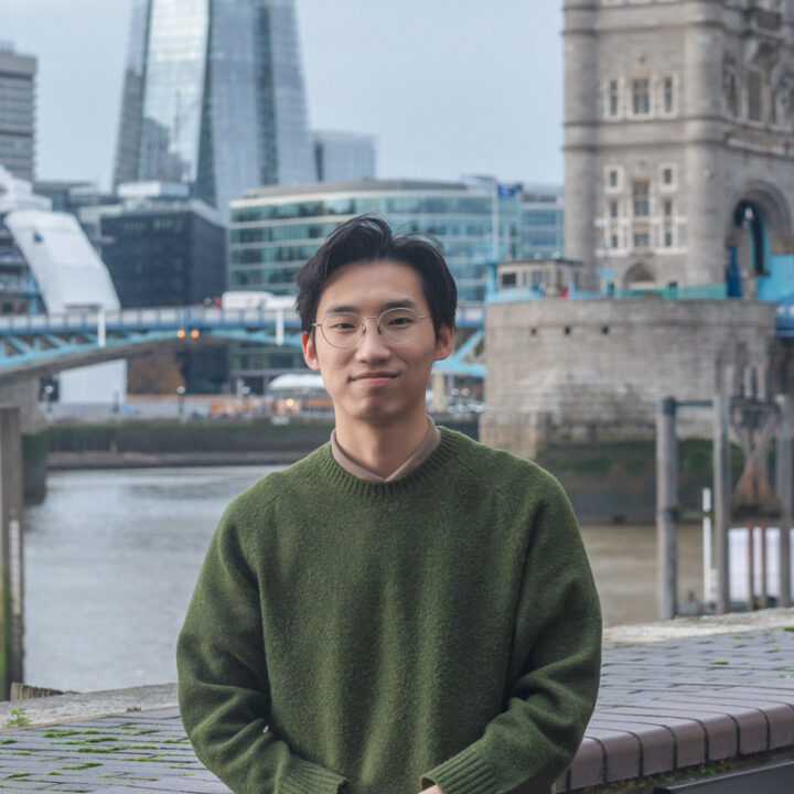 A person wearing glasses and a green sweater stands by the river with modern and historic buildings, including Tower Bridge, in the background. The scene looks overcast and urban.