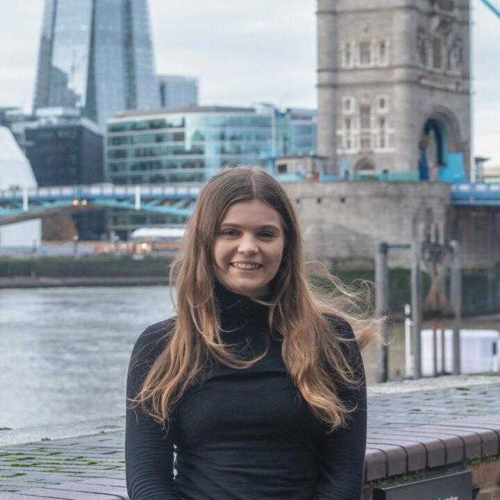 A young woman with long brown hair, wearing a black turtleneck, smiles while standing outdoors near the River Thames in London, with Tower Bridge and modern buildings visible in the background.