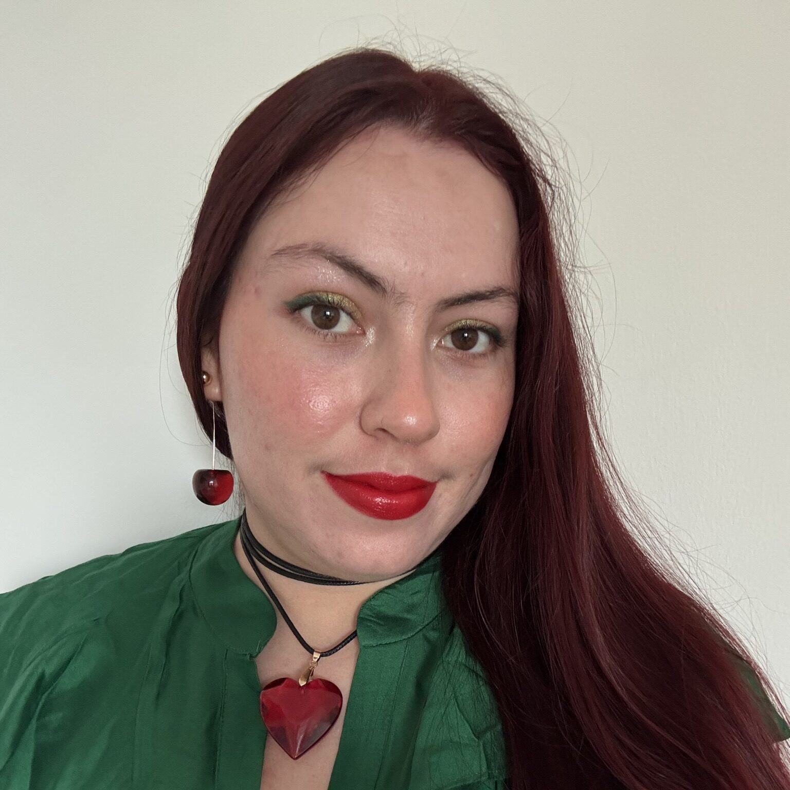 A woman with long, reddish-brown hair wears a green blouse, bold red lipstick, and matching heart-shaped red jewelry—a pendant necklace and an earring. She poses in front of a plain white background, smiling softly.