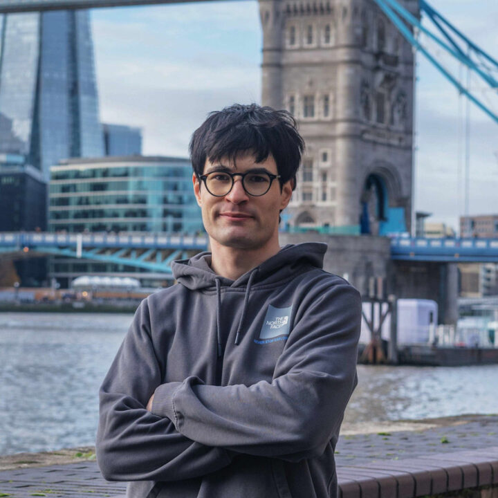 A person with dark hair and glasses, wearing a gray hoodie, stands with arms crossed by a river. Tower Bridge and modern city buildings are visible in the background.