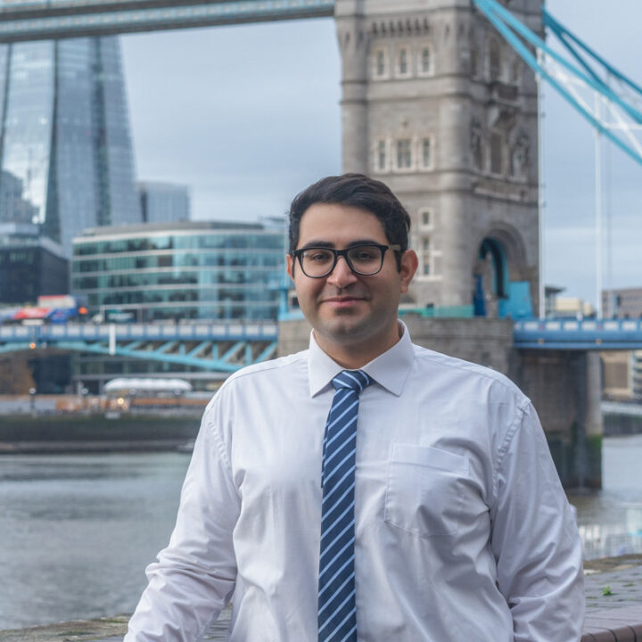 A man wearing glasses, a white shirt, and a striped blue tie stands near the Thames River with London’s Tower Bridge and modern city buildings in the background.