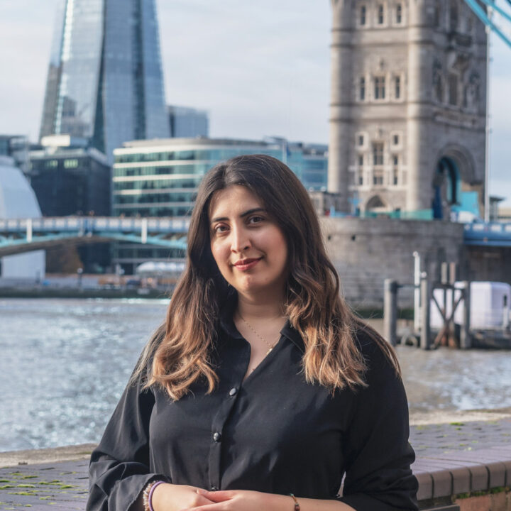 A woman with long brown hair wearing a black shirt stands by the river with modern glass buildings and the historic Tower Bridge in London in the background.