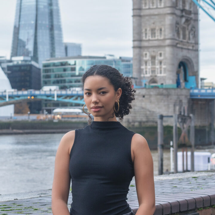 A woman with curly hair wearing a sleeveless black turtleneck top stands by a riverside with the Tower Bridge and modern buildings in the background in London.