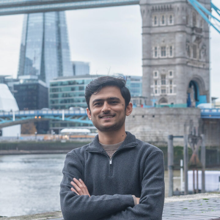A man with dark hair and a trimmed beard smiles with arms crossed near a river, with London’s Tower Bridge and modern buildings, including The Shard, in the background on a cloudy day.