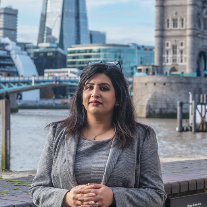 A woman in a gray blazer stands in front of a river with modern and historic buildings, including the Tower Bridge, in the background. She is facing the camera, with her hands clasped and sunglasses resting on her head.