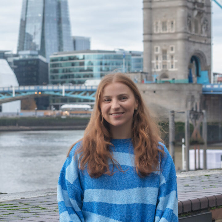 A young woman with long, wavy red hair smiles while standing by a riverside. She wears a blue and white striped sweater. Modern glass buildings and a historic stone tower are visible in the background.