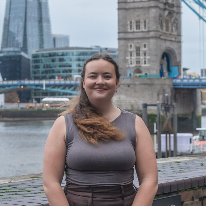 A woman with long brown hair in a ponytail smiles while standing by a river, with modern glass buildings and a stone tower bridge in the background. It appears to be a cloudy day in an urban setting.