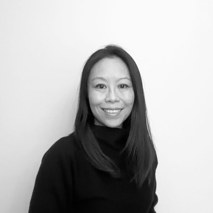 Silan Yip Levelle, with straight, long hair and a warm smile, stands against a plain, light-colored background in this black and white portrait.