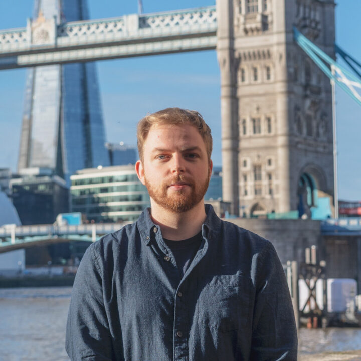 A man with light brown hair and a beard stands in front of the River Thames, with the Tower Bridge and The Shard visible in the background on a sunny day in London.