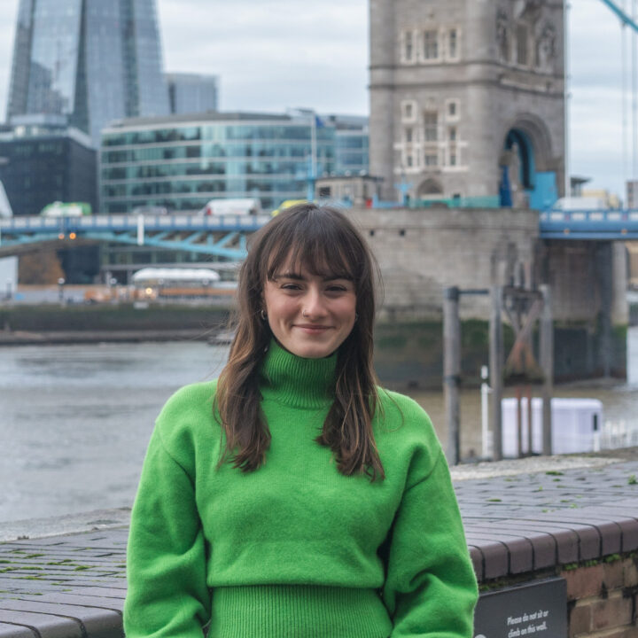 A woman wearing a bright green sweater stands smiling in front of the River Thames, with Tower Bridge and modern London buildings visible in the background on a cloudy day.