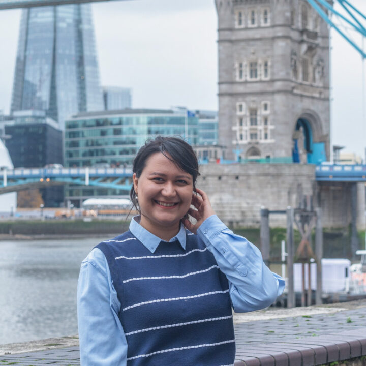 A woman smiles while standing outdoors by the Thames River with Tower Bridge and modern London buildings visible in the background on a cloudy day.