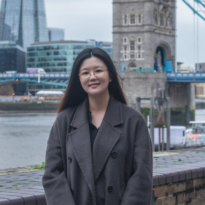 A woman with long dark hair and glasses, wearing a dark coat, stands smiling by a riverside with the Tower Bridge and modern buildings in the background.